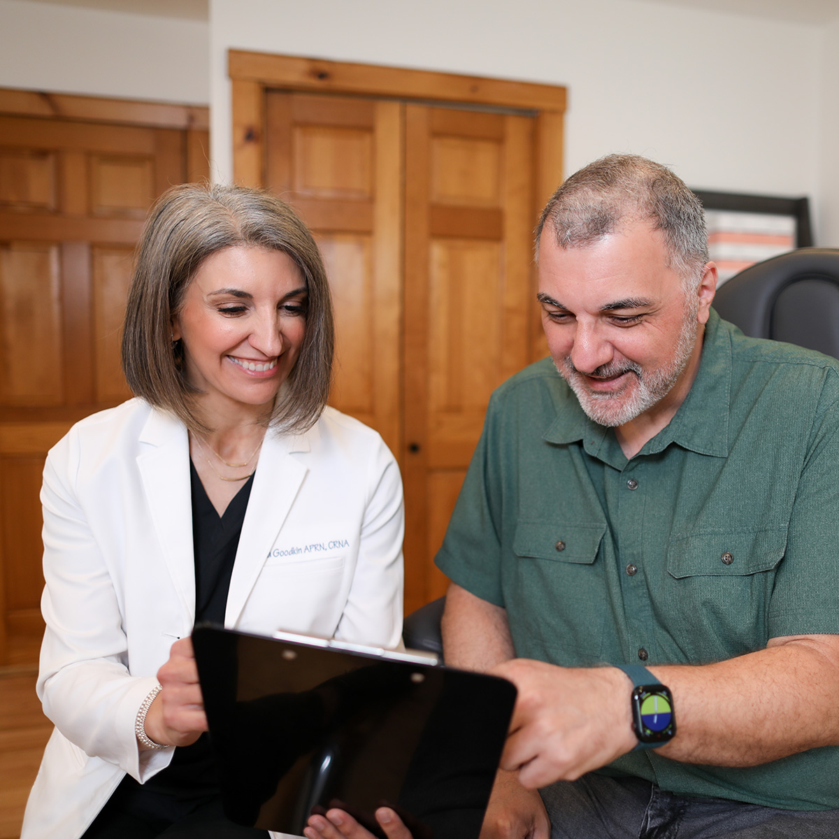 Female healthcare professional in white coat smiling and showing information on a tablet to a middle-aged man wearing a green shirt.