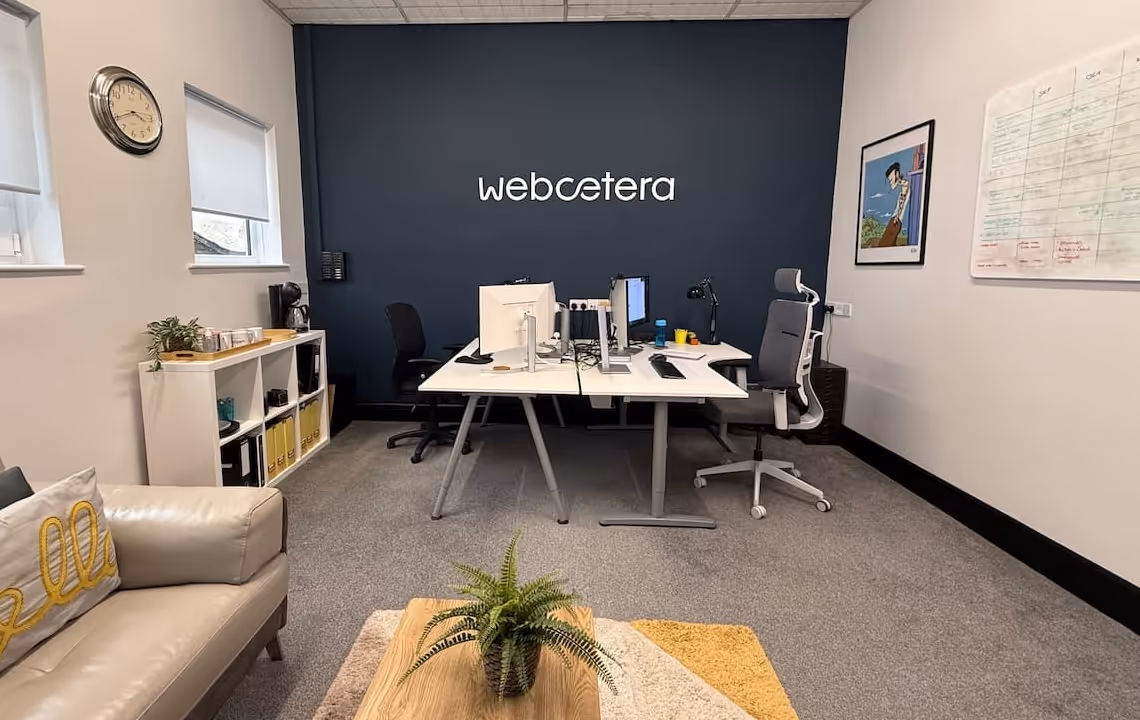 Modern office with two desks, computer monitors, ergonomic chairs, a beige sofa with a yellow 'hello' pillow, and a dark blue wall with 'webcetera' logo.