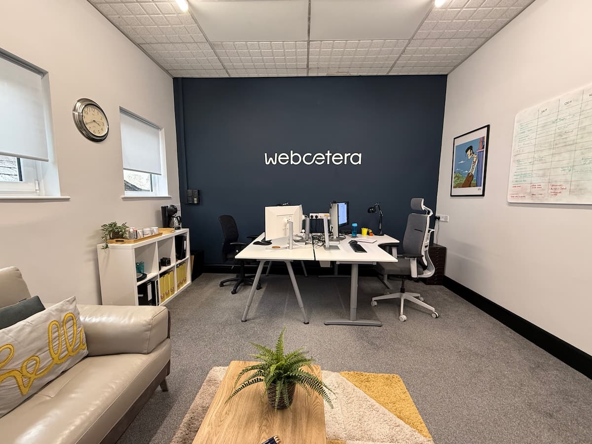 A wide-angle view of the modern Webcetera office, featuring a clean workspace with white desks, ergonomic chairs, and the Webcetera logo mounted on a navy blue feature wall.