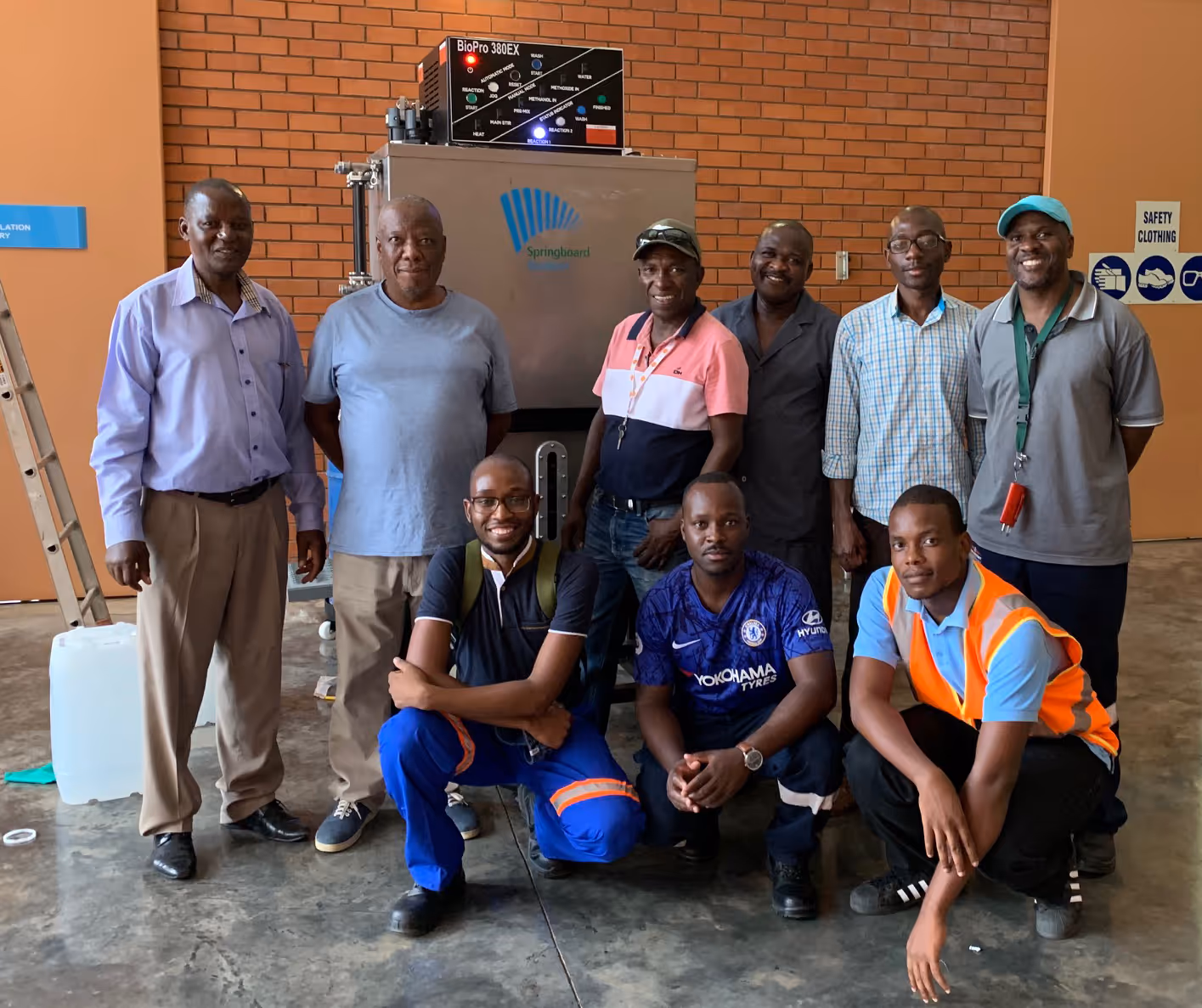 Group of nine men posing indoors in front of industrial equipment with brick wall background.