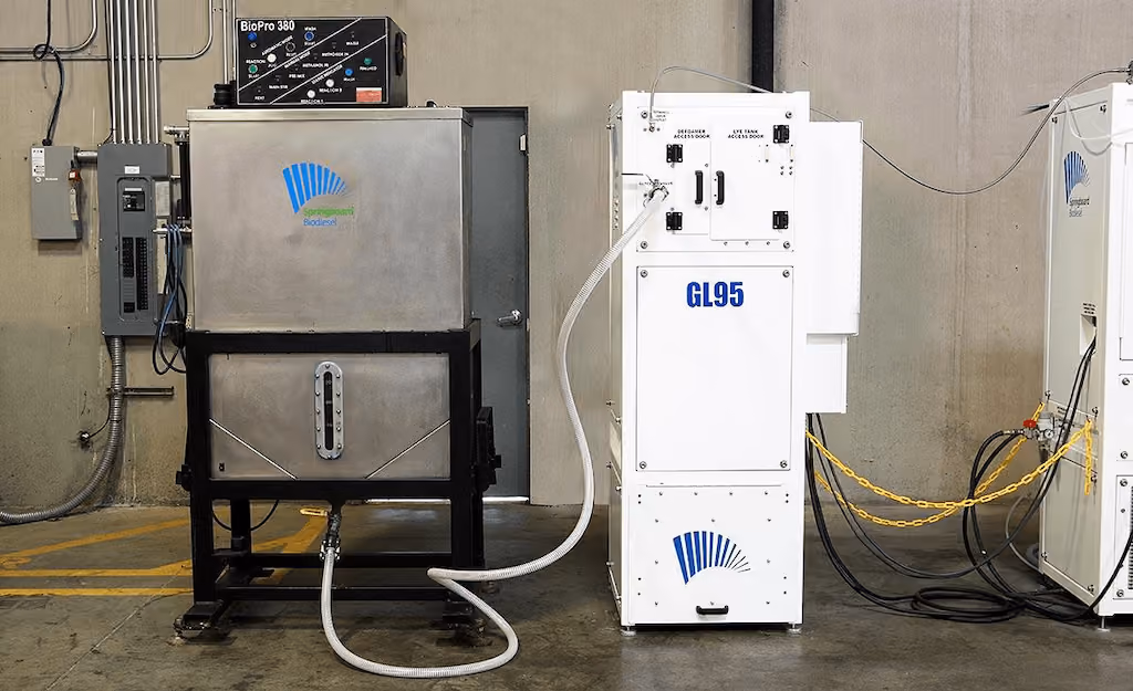 Industrial biodiesel processing equipment including stainless steel tanks labeled Biopro 380 and GL95 connected by white hoses inside a concrete room.