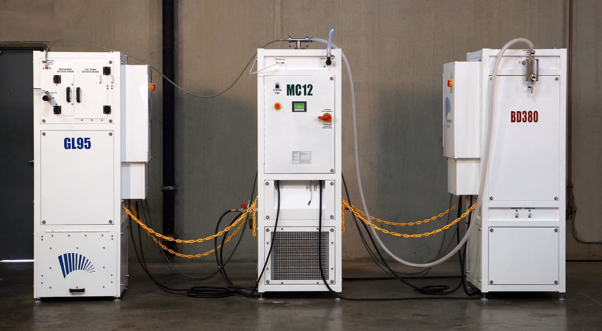Three white industrial machines labeled GL95, MC12, and BD380 connected with yellow chains and cables in a warehouse setting.