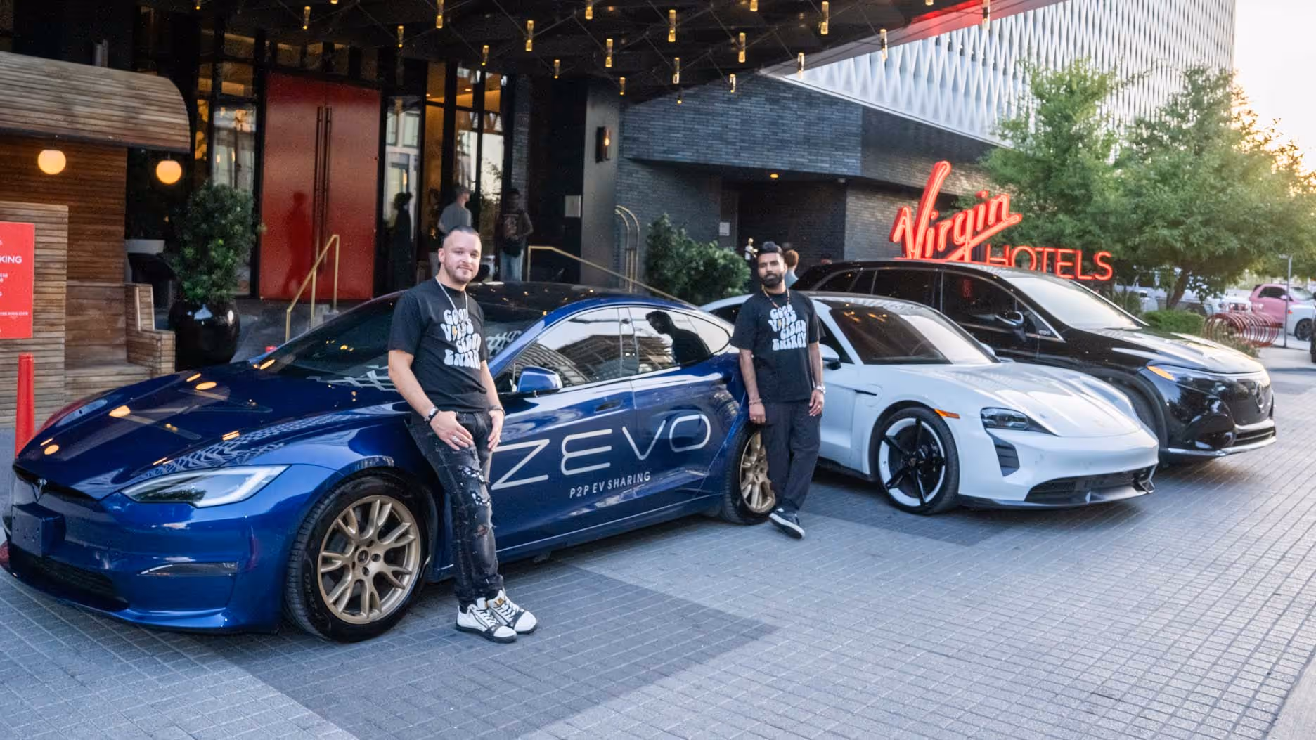 Two men in black T-shirts leaning against blue and white electric cars parked outside Virgin Hotels.