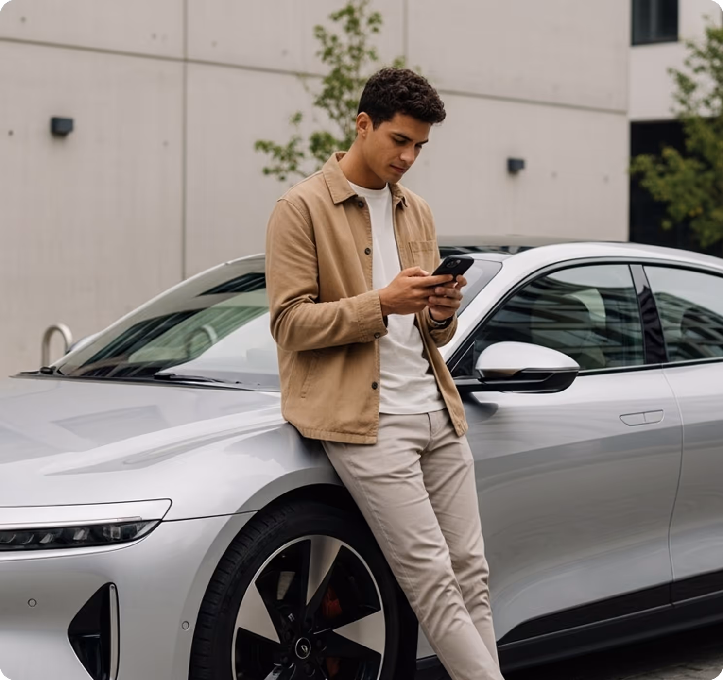 Young man in beige jacket and pants leaning against a silver car while using his smartphone.
