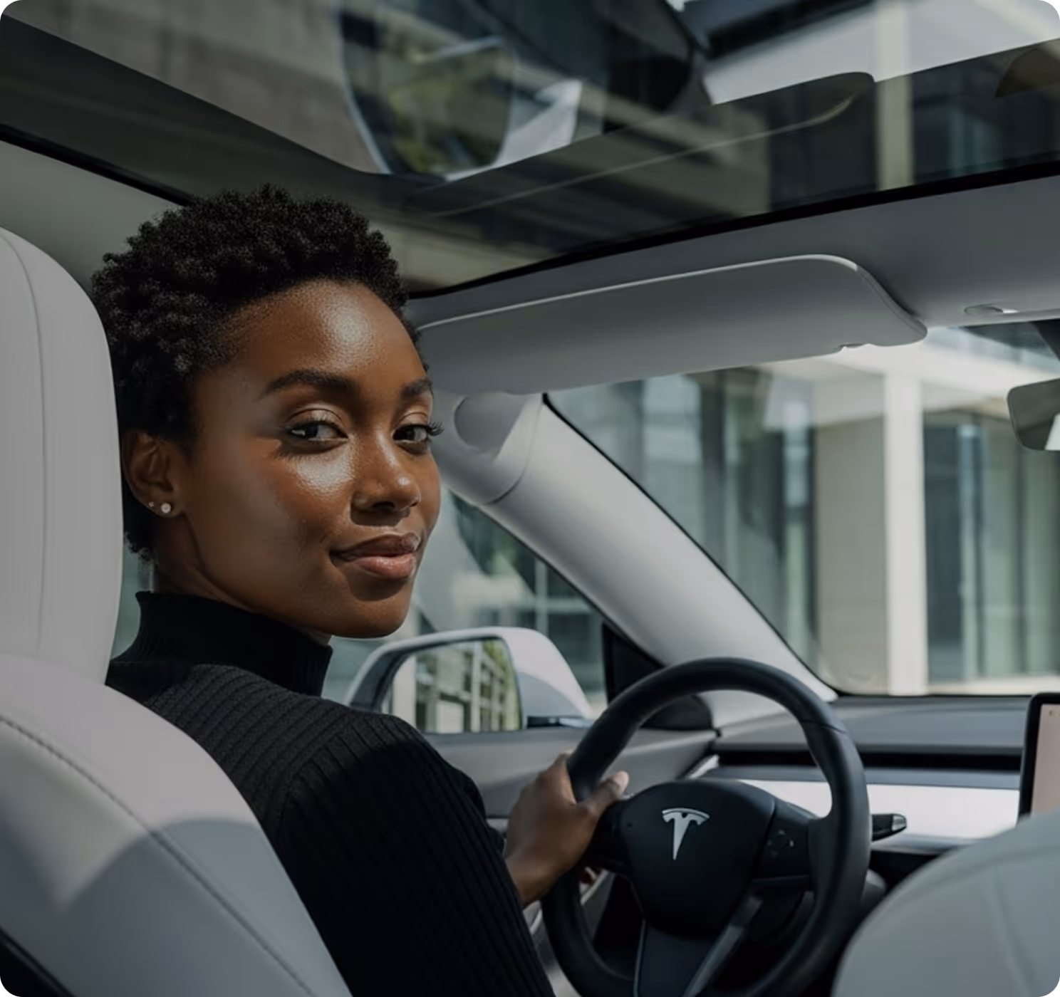 Woman with short curly hair wearing a black shirt sitting in the driver seat of a Tesla car, looking back over her shoulder.