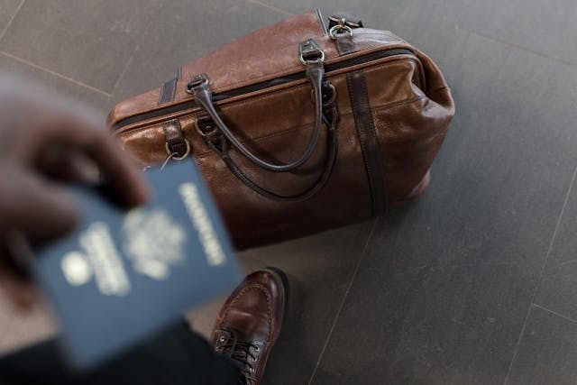 A person holds a passport next to a brown leather bag, ready for travel.