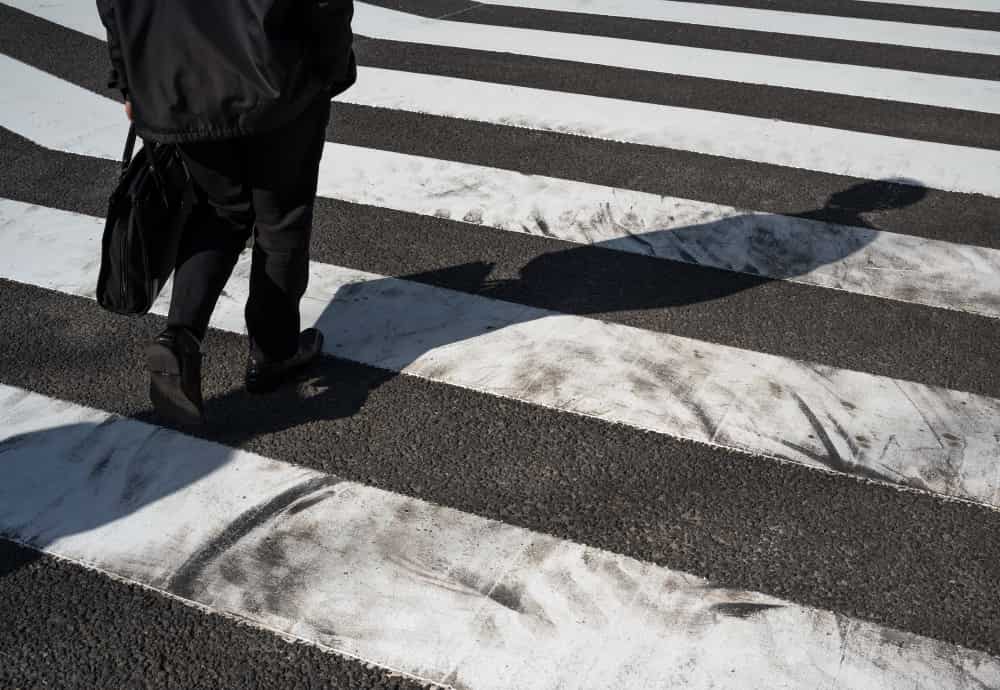 A person walks across a worn pedestrian crosswalk, casting a long shadow on the ground.