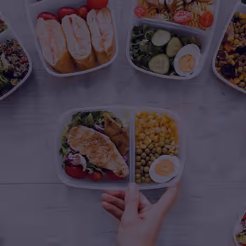 Hand holding a balanced meal container with grilled chicken salad, corn, peas, and a boiled egg.