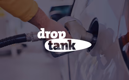 Hand holding fuel pump nozzle inserted into car's gas tank with 'drop tank' text overlay.