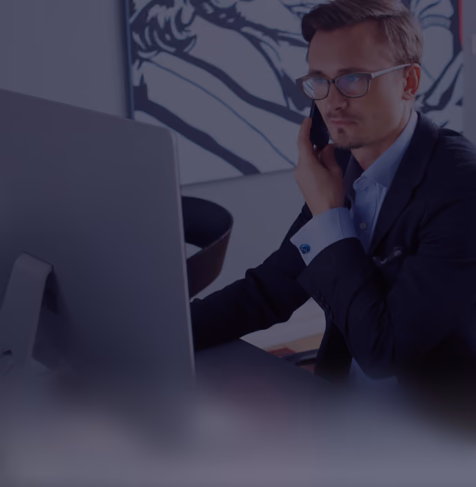 Man in glasses and suit jacket working at a computer while talking on the phone.