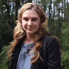 Smiling woman with long wavy blonde hair wearing a dark jacket and blue blouse standing outdoors in front of green trees.