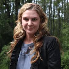 Smiling woman with long wavy blonde hair wearing a dark jacket and blue blouse standing outdoors in front of green trees.