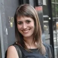 Smiling woman with shoulder-length brown hair wearing a black sleeveless top in an urban setting.