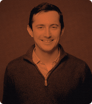 Portrait of a smiling man with short dark hair wearing a dark quarter-zip sweater over a patterned shirt.