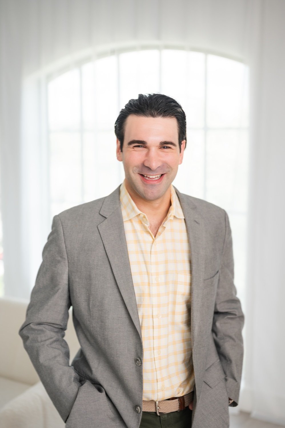 Smiling man in a gray blazer and yellow plaid shirt standing indoors in front of a window with sheer curtains.