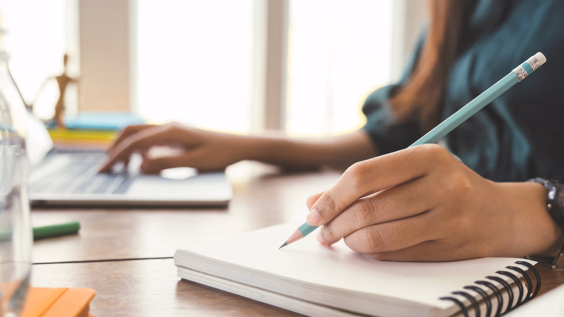 Woman at desk writing on notebook and using laptop