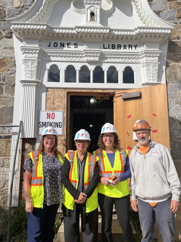 four people in hard hats and yellow vests stand in front of the entrance to the Jones Library
