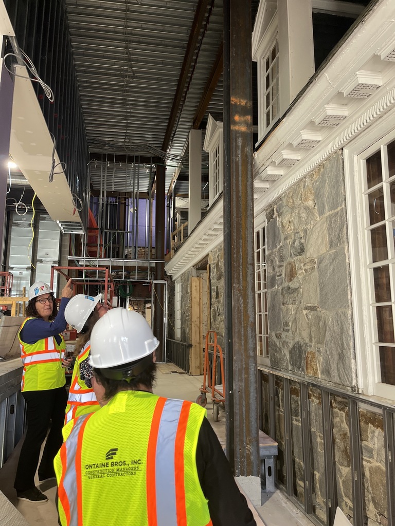 three people in hard hats and yellow vests look up at the now-interior wall of the exterior of the Jones Library, with stonework and gabled windows