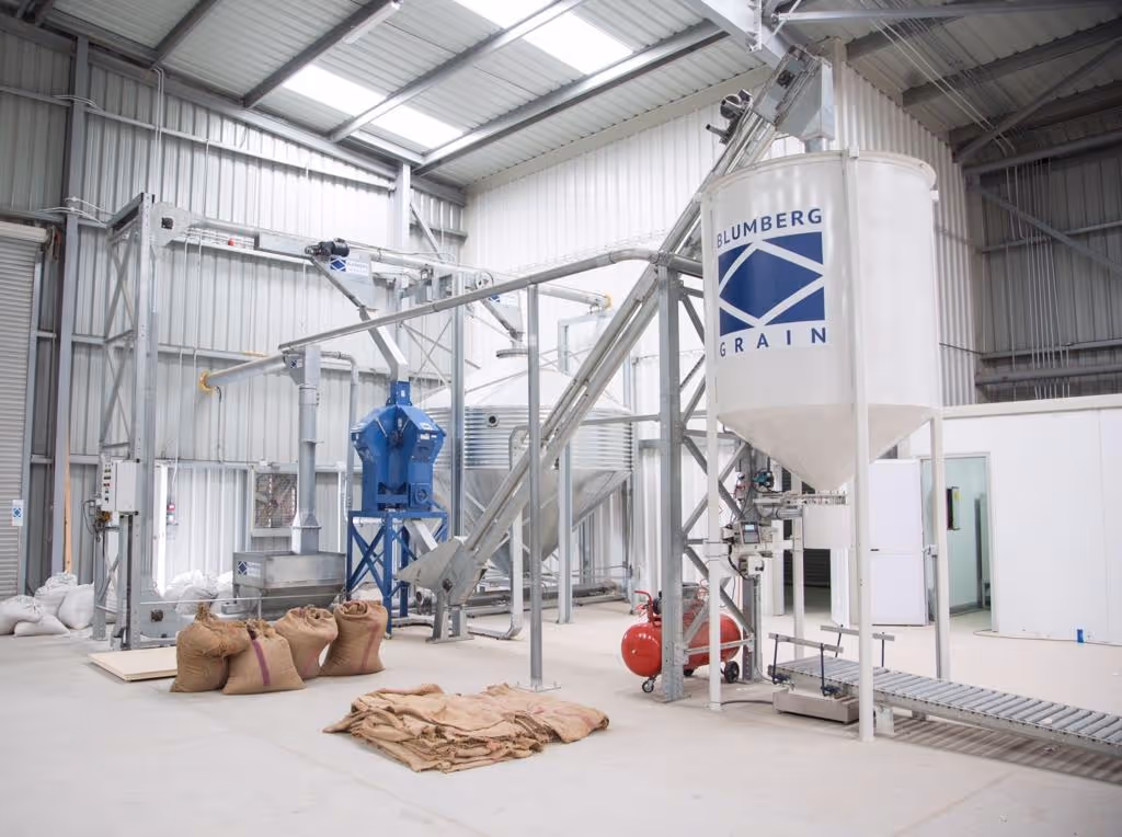 Industrial grain processing facility interior with Blumberg Grain silo, conveyor system, blue machinery, and burlap sacks on the floor.