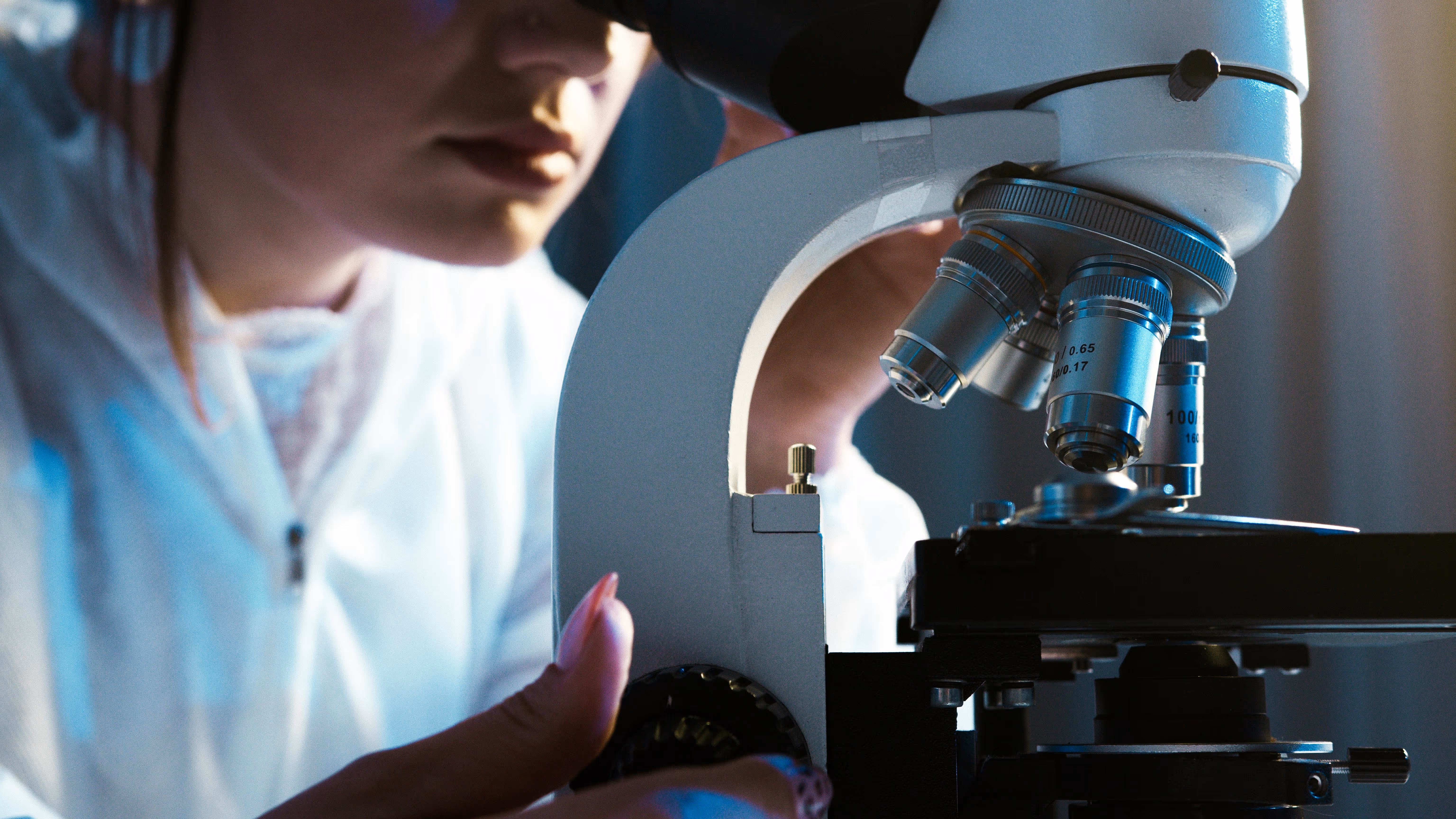 Person adjusting the focus of a microscope while looking through the eyepiece in a laboratory setting.