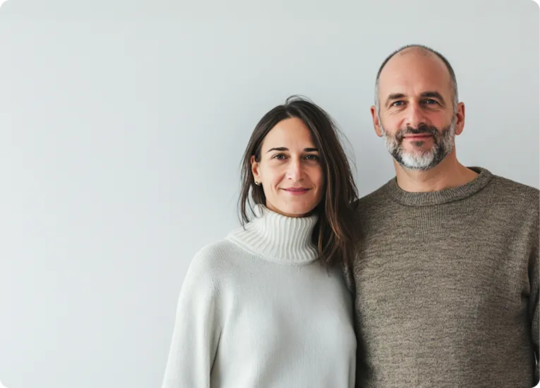 Smiling man with a beard and woman with long brown hair, both wearing sweaters, standing side by side against a plain light background.