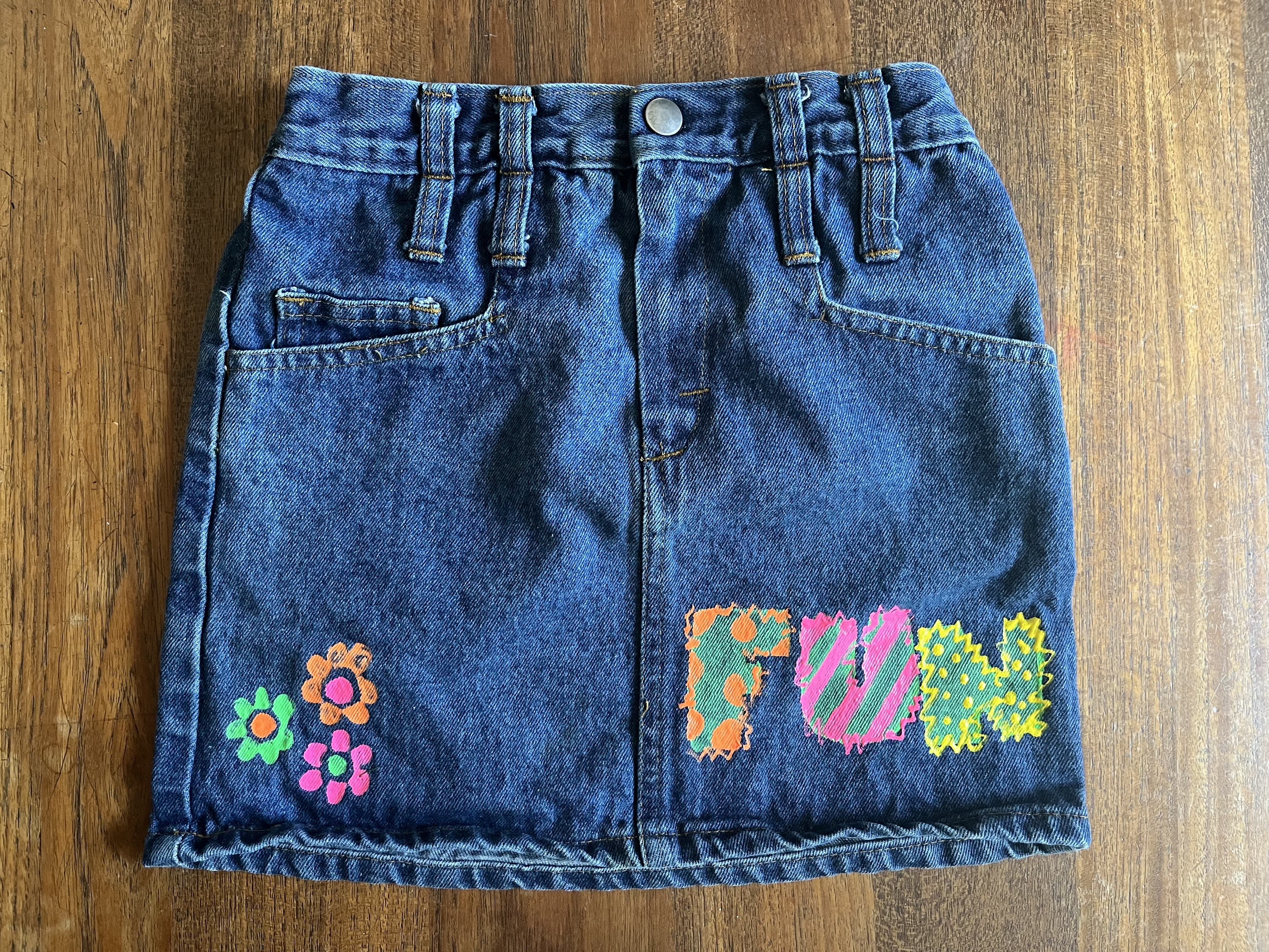 A jean skirt with "FUN" and several flowers painted in bright orange, pink, and blue tones by school-aged Precious