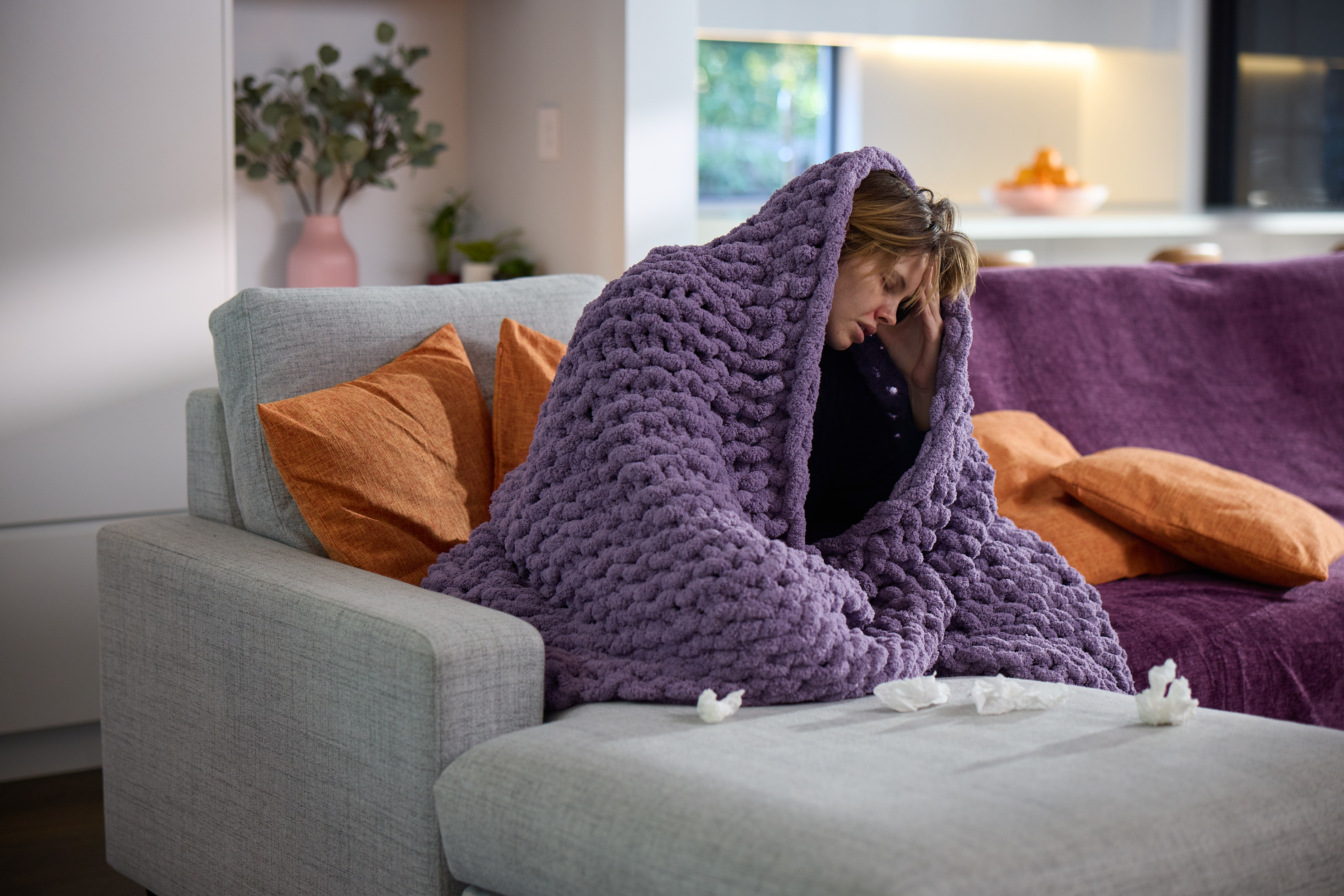 Unwell woman wrapped in a purple blanket on the couch, about to request a sick certificate online