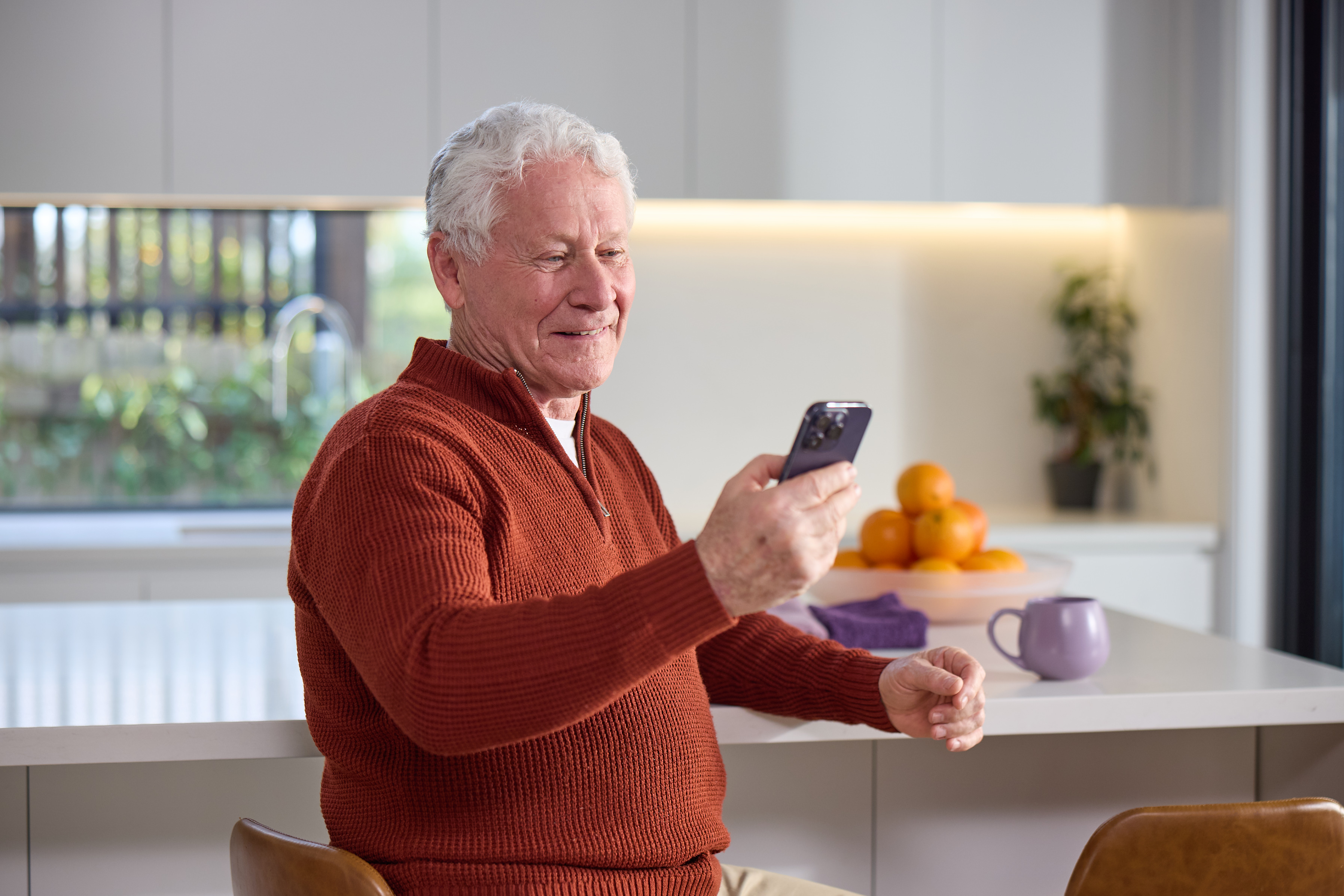 Smiling older man in a red jumper at his kitchen bench, checking his phone about a heart condition