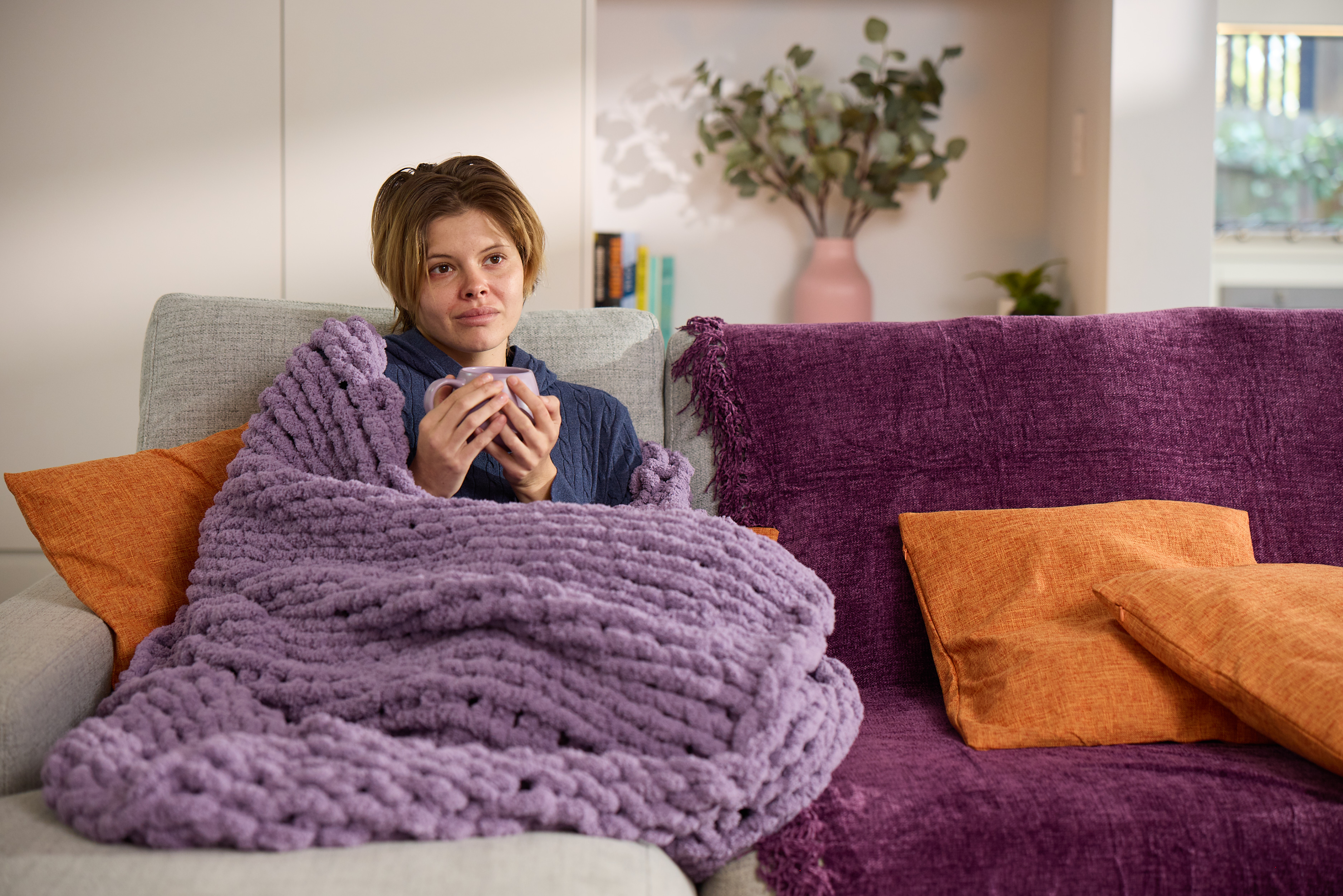Young woman wrapped in a purple knitted blanket on the couch, holding a mug while exploring depression treatment options