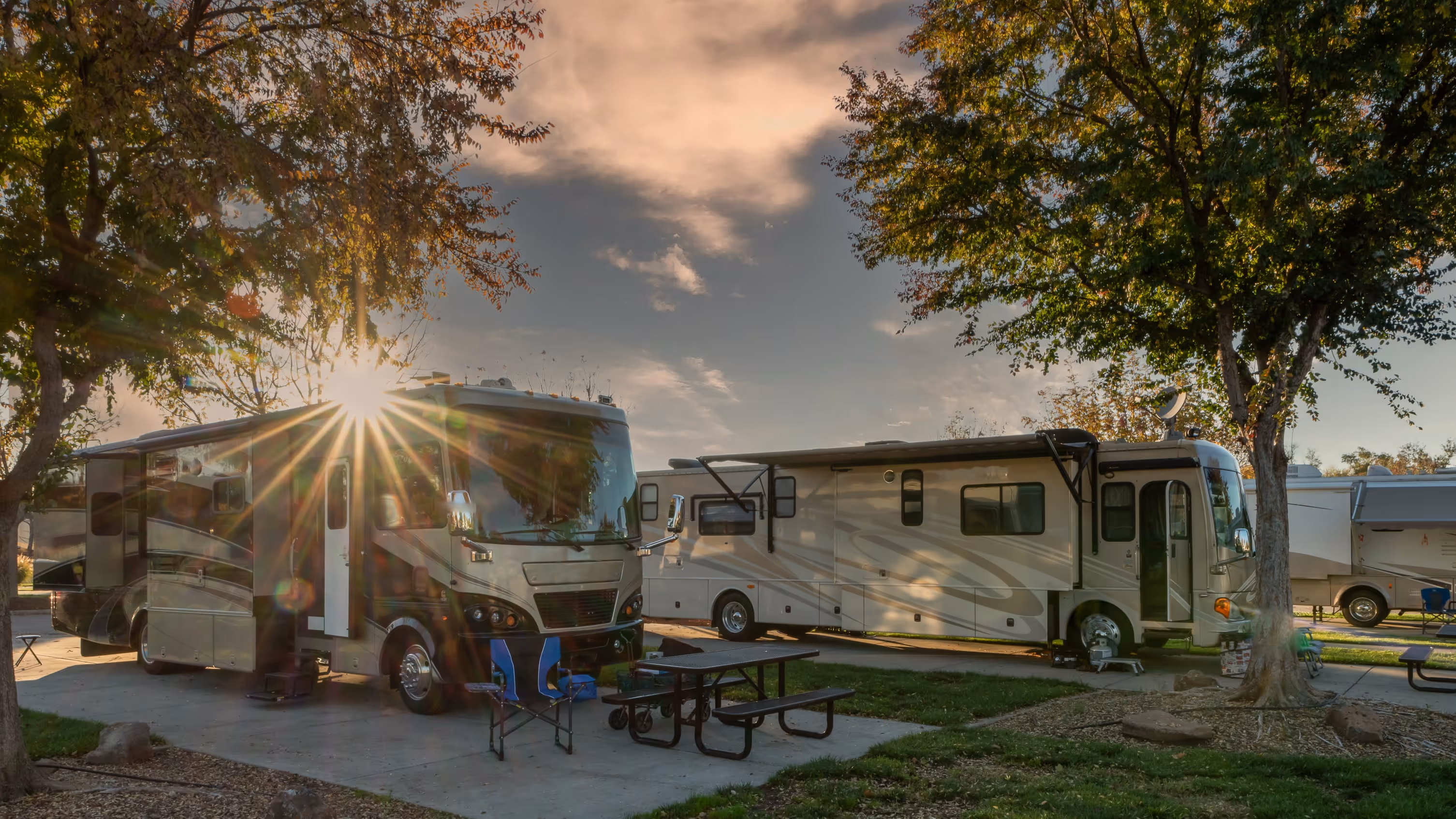 Two large recreational vehicles parked at a campsite with picnic tables and trees during sunset.