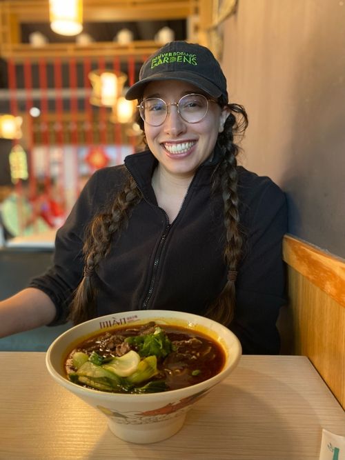 A picture of woman with long dark, braided hair, glasses, and a baseball cap. She is sitting in front of a bowl of noodles.