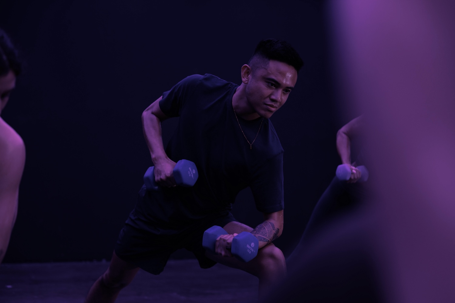 Person in black workout clothes lifting blue dumbbells in purple light