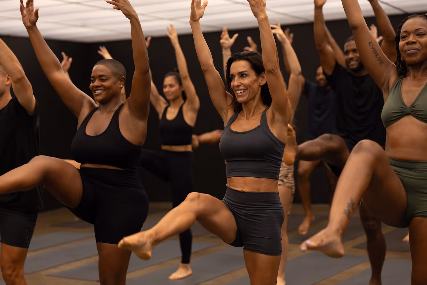 Diverse group of people in athletic wear performing a barre workout with arms raised and legs lifted.