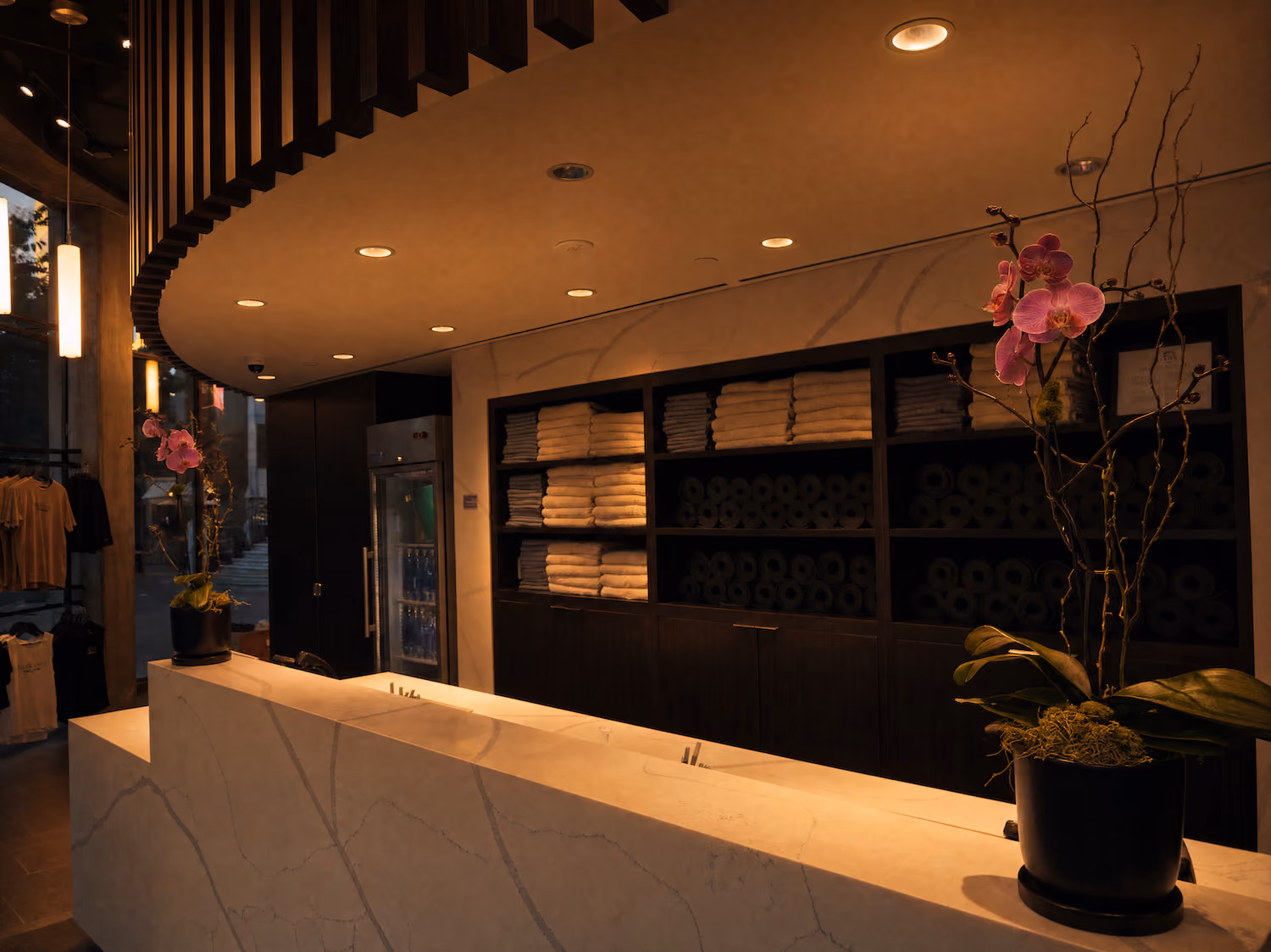 Modern spa reception with white marble desk, towel shelves, pink orchids, warm lighting, and elegant decor.