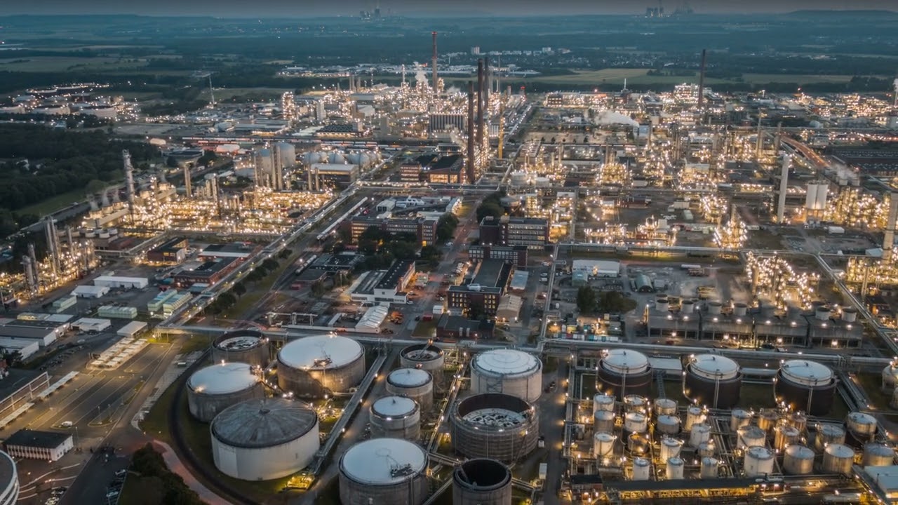 Aerial view of a large industrial refinery complex at dusk with numerous storage tanks and illuminated pipes and buildings.