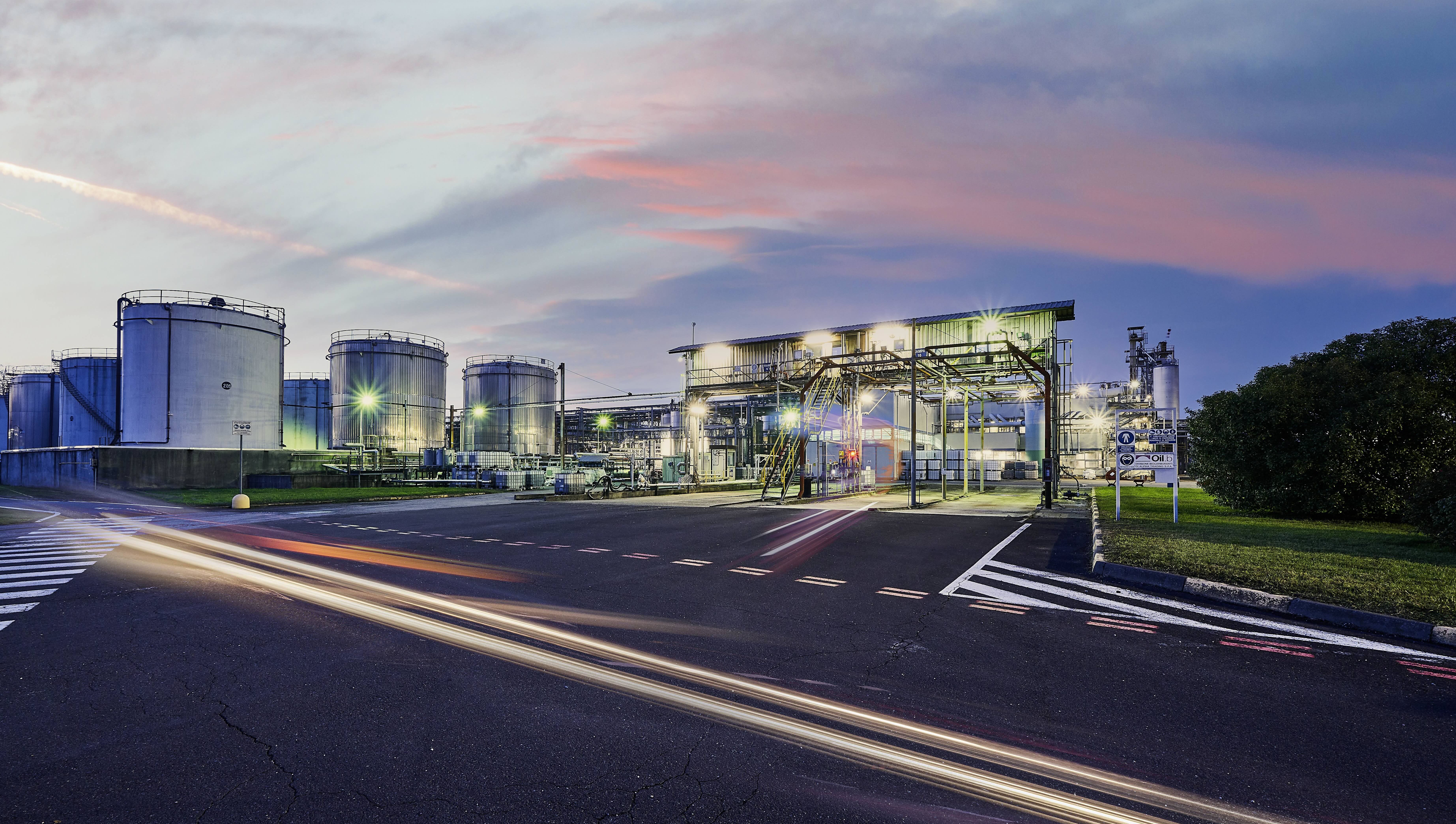 Industrial facility with large storage tanks and bright lights at dusk under a colorful sky.