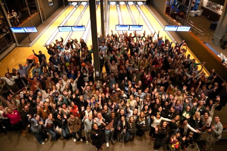 Large group of people waving and smiling at a bowling alley with multiple lanes in the background.