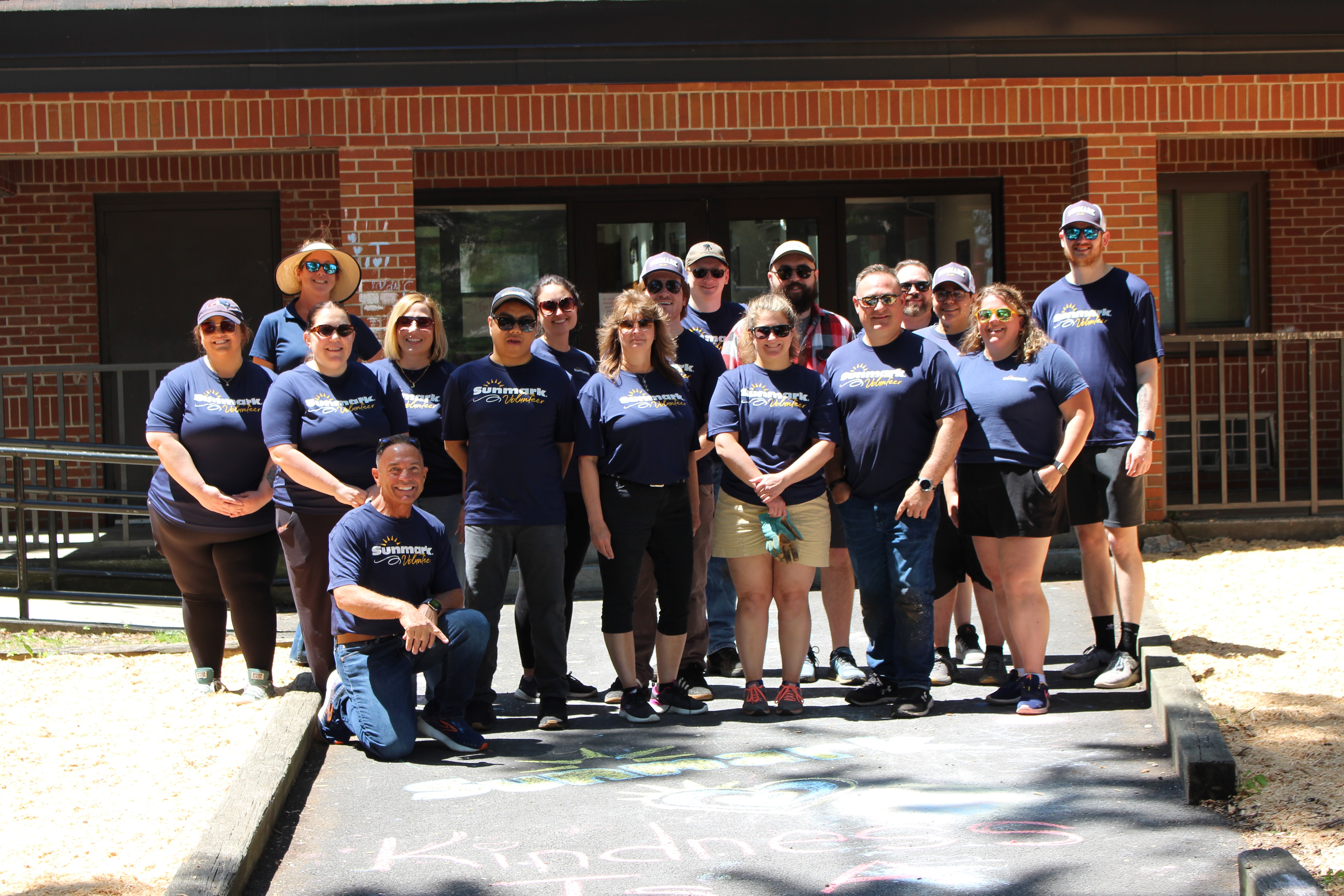 Group of smiling volunteers in matching Sunmark Volunteer shirts posing outside a brick building.