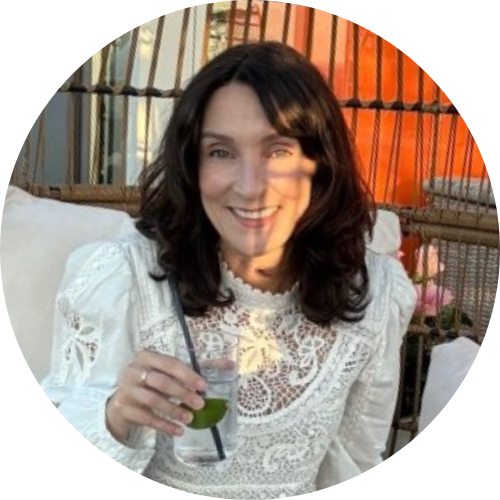 Smiling woman with dark hair wearing a white lace top and holding a drink while seated outdoors.