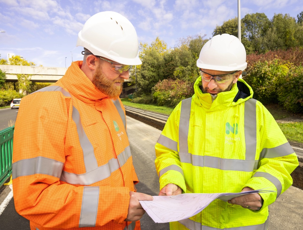 Two construction workers wearing hard hats and high-visibility jackets review a set of plans together at a roadside work site.