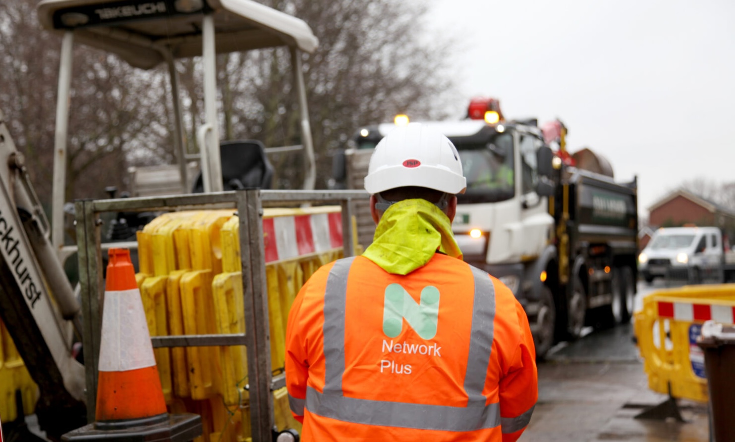 Construction worker in high-visibility gear and a hard hat stands at a roadside work site with utility vehicles and equipment.