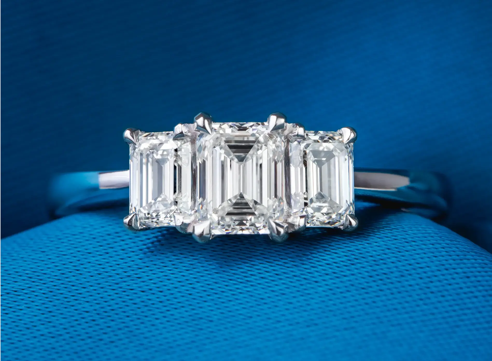 Silver ring with three emerald-cut diamonds displayed on blue textured fabric.