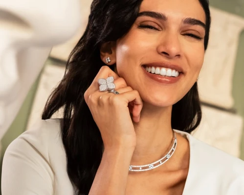 Smiling woman with long dark hair wearing sparkling diamond jewelry including a ring, necklace, and earrings.