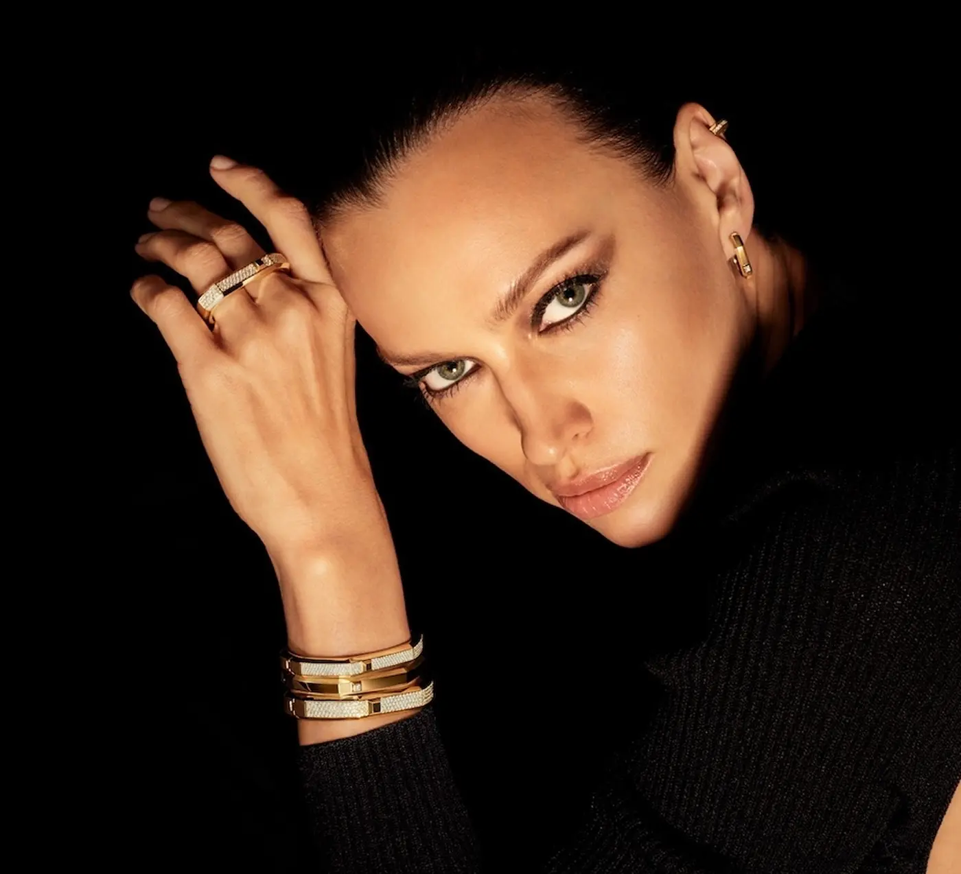 Close-up of a woman with green eyes wearing gold and white bracelets, rings, and earrings against a black background.