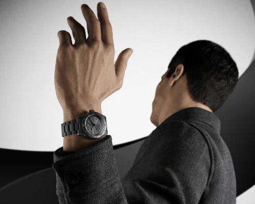 Man in gray coat raising hand, showing a black wristwatch against a curved light background.