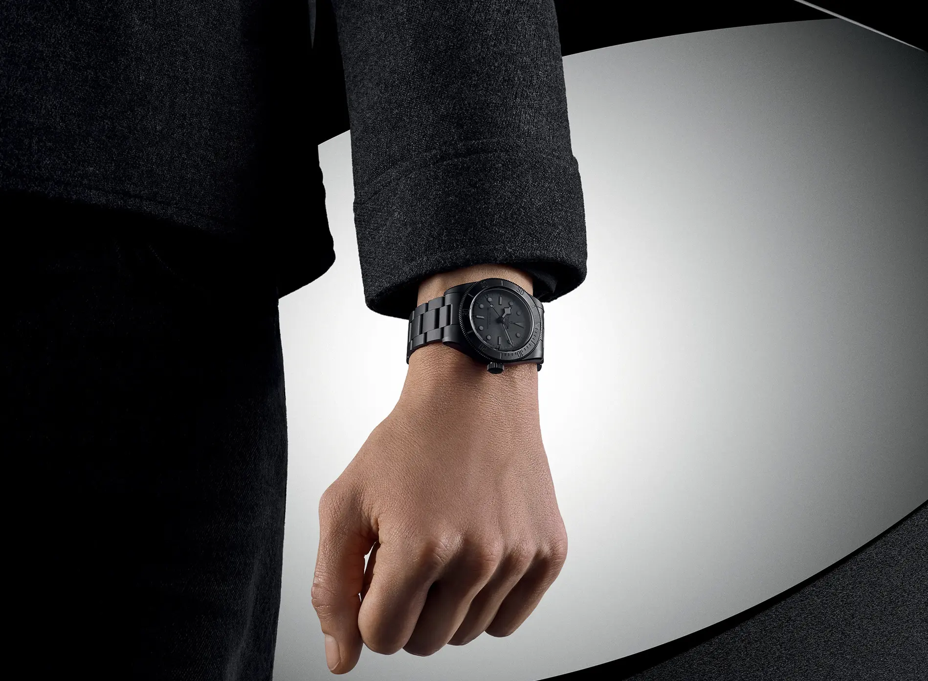 A person's hand wearing a black metal wristwatch with a dark dial and bracelet against a gray background.