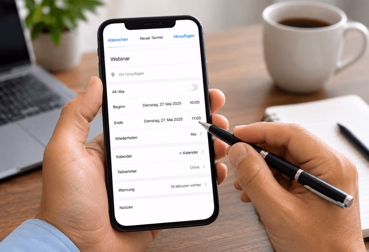 Smartphone zeigt Online-Kalender mit Webinar-Details, gehalten von einer Hand mit Stift neben Laptop und Kaffeetasse.