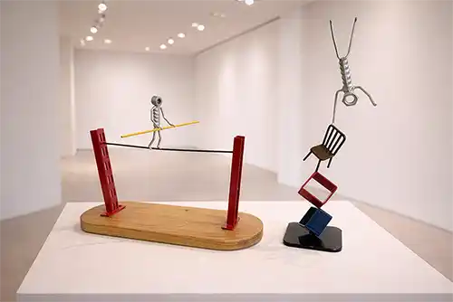Two recycled metal sculpture. One is a tightrope walker, the other is an acrobat balanced with one hand on a chair and two square hollow structures