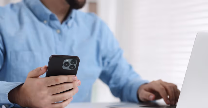 young man using smartphone while working with laptop at white table in office
