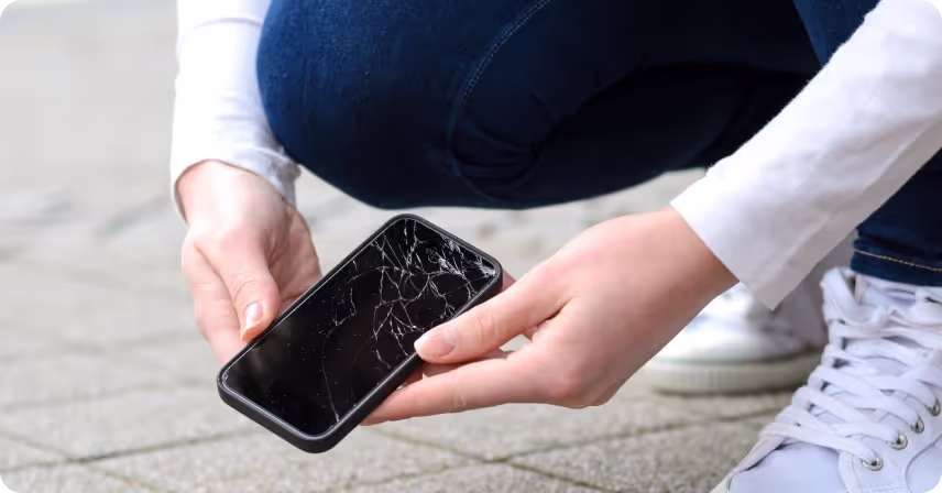 a person wearing jeans and a white shirt is crouching down to pick up a smartphone with a severely cracked screen